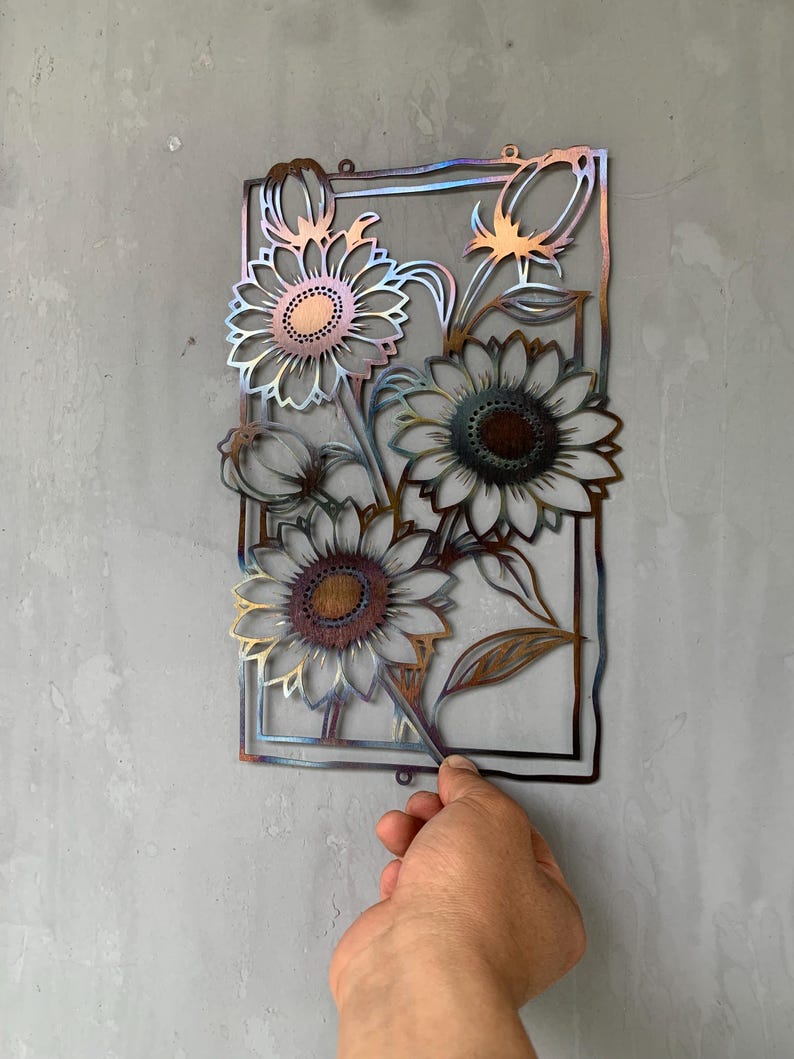 May include: Metal wall art featuring sunflowers in a rectangular frame. The sunflowers have detailed petals and textured centers, with a mix of bronze, gold, and blue hues. The piece is designed to be mounted on a wall.