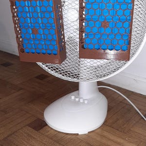Könnte beinhalten: Ein weißer elektrischer Ventilator mit zwei braunen, 3D-gedruckten Halterungen mit Sechseckmuster, die am Frontgitter befestigt sind. Jede Halterung enthält ein rechteckiges blaues Kühlelement, um den Luftstrom des Ventilators zu kühlen.