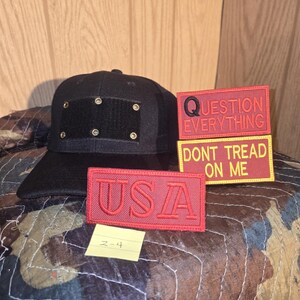 May include: Black baseball cap with a flat peak and a square patch with brass eyelets. Includes three red embroidered patches with the text "USA", "QUESTION EVERYTHING", and "DONT TREAD ON ME".