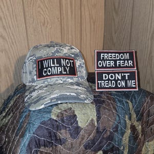 May include: A camouflage baseball cap with the text "I WILL NOT COMPLY" in a red-bordered black rectangle. Two patches read "FREEDOM OVER FEAR" and "DON'T TREAD ON ME". The items are displayed on a camouflage patterned surface.