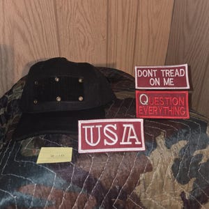May include: Black baseball cap with a square patch and metal rivets. Three embroidered patches with the phrases "DONT TREAD ON ME", "QUESTION EVERYTHING", and "USA" in red and white. The items are on a camouflage patterned surface.