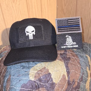 May include: Black baseball cap with a white skull patch. Two additional patches feature an American flag with a blue line and a coiled snake with the text "DON'T TREAD ON ME". The items are displayed on a camouflage patterned surface.