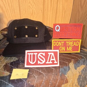 May include: A black baseball cap with a square patch and gold rivets. Three rectangular patches with text: "QUESTION EVERYTHING", "DONT TREAD ON ME", and "USA" in red and white. A yellow sticky note is also present.