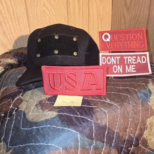 May include: Black baseball cap with a square patch and eyelets. Includes three red patches with white text: "QUESTION EVERYTHING", "DONT TREAD ON ME", and "USA". The patches are rectangular and have embroidered lettering.