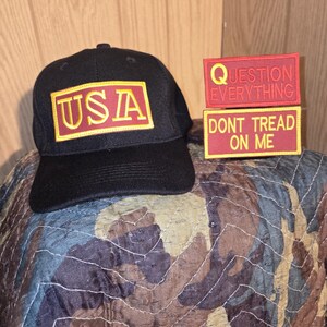 May include: Black baseball cap with a rectangular patch reading "USA" in yellow on red. Two rectangular patches with text: "QUESTION EVERYTHING" and "DONT TREAD ON ME" in yellow on red. The items are on a camouflage patterned surface.
