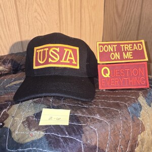 May include: A black baseball cap with a rectangular patch displaying "USA" in gold lettering on a red background. Two red rectangular patches with gold text are also visible, one reading "DONT TREAD ON ME" and the other "QUESTION EVERYTHING".