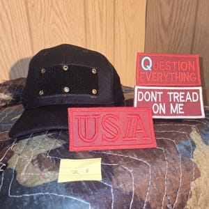 May include: A black baseball cap with a rectangular patch and brass-coloured rivets. Three red rectangular patches with white text that reads "QUESTION EVERYTHING", "DONT TREAD ON ME", and "USA".