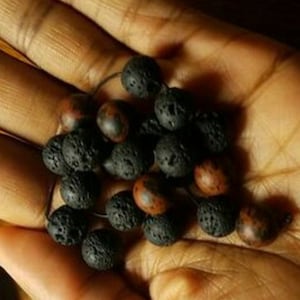 May include: A close-up of a hand holding a strand of black lava rock beads with brown beads interspersed throughout.
