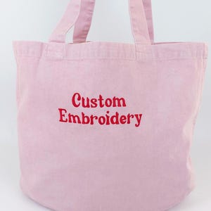 Può includere: Una borsa tote rosa chiaro con manici coordinati. Le parole "Custom Embroidery" sono ricamate in rosso sul davanti. La borsa è realizzata in un tessuto morbido ed è progettata per trasportare oggetti.