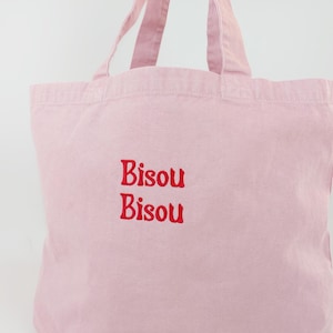 May include: A light pink tote bag with two handles. The bag has the embroidered text "Bisou Bisou" in red. The bag is made of a soft, durable fabric and is suitable for carrying various items.