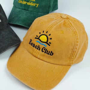 Peut inclure: Trois casquettes de baseball avec une finition délavée. Une casquette gris anthracite porte le texte brodé blanc Girl Dad. Une casquette vert foncé porte le texte brodé jaune Custom Embroidery. Une casquette jaune moutarde porte un motif soleil et le texte Beach Club.