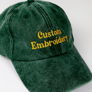Pode incluir: Um boné de beisebol verde-escuro com uma textura lavada e desgastada. A parte frontal apresenta o texto Custom Embroidery bordado em fio amarelo. O boné tem uma aba curva, um botão no topo central e ilhós costurados para ventilação.