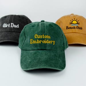 Op de afbeelding: Drie baseballpetten in verschillende kleuren. Eén is donkergrijs met wit geborduurd "Girl Dad". Een andere is groen met geel "Custom Embroidery". De derde is bruin met een zon en "Beach Club" in zwart.