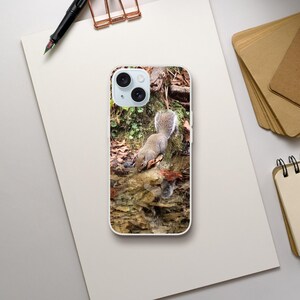 Könnte beinhalten: Ein hellgraues Eichhörnchen mit buschigem Schwanz trinkt aus einem kleinen Bach. Das Eichhörnchen befindet sich an einem felsigen Ufer mit abgefallenen Blättern. Die Szene wird auf einer Handyhülle mit weißem Rand festgehalten. Ein Stift und Papier befinden sich im Hintergrund.