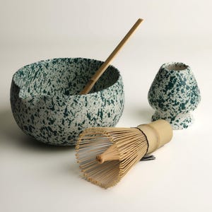 Op de afbeelding: Een matcha-theeset met een gespikkelde keramische kom, een garde, een bamboelepel en een keramische theekop. De kom en kop hebben een witte basis met donkergroene spetters. De garde is gemaakt van bamboe.