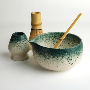 Könnte beinhalten: Ein Matcha-Teeset mit einer gesprenkelten grün-weißen Keramikschale mit Ausguss, einem Bambusbesen, einem Bambuslöffel und einer kleinen Keramik-Teeschale. Das Set ist für die Zubereitung von Matcha-Tee konzipiert.