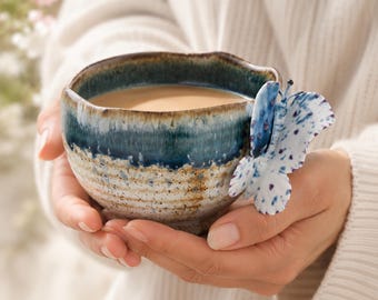 May include: A ceramic bowl with a blue and brown glaze, filled with a light brown liquid. A decorative butterfly with blue and purple spots is attached to the bowl. The bowl is held in both hands.