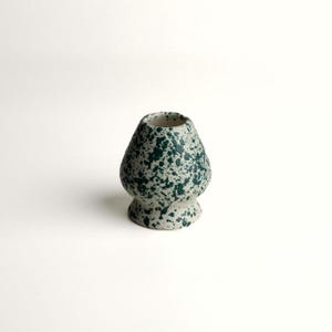 May include: A ceramic matcha whisk holder featuring a speckled pattern with dark green paint splatters on a light cream-colored base. The object has a rounded, bulbous body that tapers toward the top opening and flares at the base.