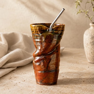 Könnte beinhalten: Ein handgefertigter Keramikbecher mit einem gedrehten Design und einem Edelstahlstrohhalm. Der Becher hat eine braune, orangefarbene und gelbe Glasur. Die einzigartige Form und die Farben machen ihn zu einem stilvollen Trinkgefäß.