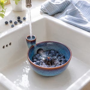 Puede incluir: Un colador de bayas redondo de cerámica esmaltada en azul y marrón con un embudo integrado para la entrada de agua. El cuenco cuenta con un cono central elevado y un pico lateral, lleno de arándanos frescos siendo enjuagados bajo un grifo.