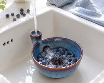 May include: A round, blue and brown glazed ceramic berry colander with a built-in funnel for water intake. The bowl features a central raised cone and a side spout, currently filled with fresh blueberries being rinsed under a kitchen tap.