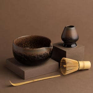 May include: A ceramic matcha bowl with a dark brown and black speckled glaze, a matching ceramic whisk holder, a light-coloured bamboo whisk with thin tines, and a long, slender bamboo scoop, all arranged on a solid brown surface.