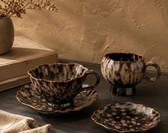 May include: Two ceramic teacups and saucers with a speckled brown and cream pattern. One cup sits on a saucer, the other cup is on a pedestal. A third saucer is also visible. The cups and saucers have a scalloped edge.