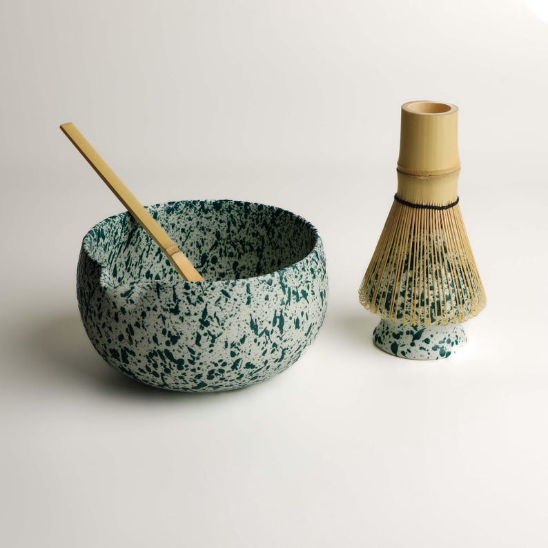 May include: A white ceramic bowl with a dark green speckled pattern, featuring a small pouring spout and a light brown bamboo whisk resting inside. Beside it stands a matching bamboo whisk holder with the same green and white speckled finish.