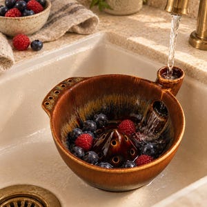 May include: A brown ceramic berry colander bowl with a central cone and a side spout, featuring a glossy glaze with dark drip accents. The bowl contains fresh raspberries and blueberries being rinsed under a stream of water from a tap.