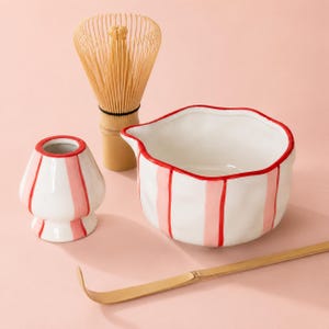 Op de afbeelding: Een keramische matchakom en een bijpassende gardehouder, beide met witte oppervlakken met verticale roze strepen en rode randen. Een bamboe matchagarde en een lange, gebogen bamboe theeschep zijn naast de keramische onderdelen geplaatst.