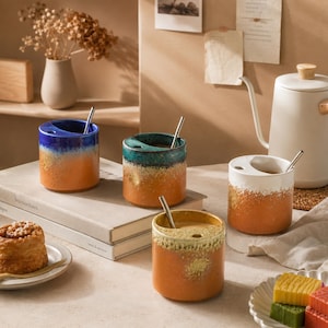 May include: Four ceramic mugs with a gradient color scheme, featuring blue, teal, white, and yellow hues over orange. Each mug has a unique design and includes a silver-colored straw. The mugs are cylindrical and appear to be handmade.