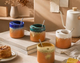 May include: Four ceramic mugs with a gradient color scheme, featuring blue, teal, white, and yellow hues over orange. Each mug has a unique design and includes a silver-colored straw. The mugs are cylindrical and appear to be handmade.