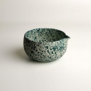 May include: A round ceramic mixing bowl with a pour spout, featuring a cream-colored base with a dark green speckled paint splatter pattern covering the entire exterior and interior surface of the vessel, creating a distinct textured appearance.