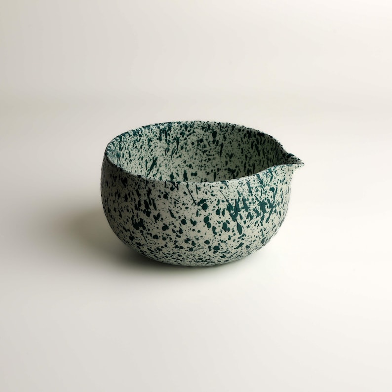 May include: A round ceramic mixing bowl with a pour spout, featuring a cream-colored base with a dark green speckled paint splatter pattern covering the entire exterior and interior surface of the vessel, creating a distinct textured appearance.
