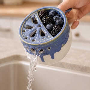 Pode incluir: Uma tigela escorredor de cerâmica para frutas vermelhas com base branca e seção superior esmaltada em azul com um padrão de recortes decorativos. A tigela está cheia de amoras e mirtilos frescos, com água escorrendo pelos orifícios.