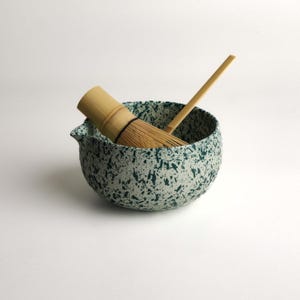May include: A ceramic matcha bowl with a white base and dark green speckled pattern, featuring a small pouring spout. Inside the bowl rests a light brown bamboo matcha whisk and a long, thin bamboo scoop, both used for preparing traditional tea.