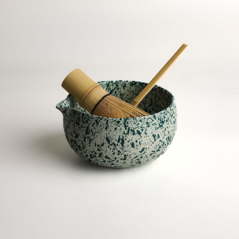 May include: A ceramic matcha bowl with a white base and dark green speckled pattern, featuring a small pouring spout. Inside the bowl rests a light brown bamboo matcha whisk and a long, thin bamboo scoop, both used for preparing traditional tea.