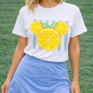 May include: White t-shirt featuring a lemon design. The graphic includes three yellow lemon slices with green leaves, set against a backdrop of blue and white stripes. The person is wearing a light blue skirt.