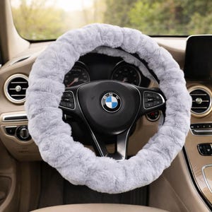 Könnte beinhalten: Ein hellgrauer, flauschiger Lenkradbezug auf einem schwarzen Lenkrad. Der Bezug besteht aus einem weichen, plüschigen Material. Das Lenkrad hat das BMW-Logo in der Mitte. Der Innenraum des Autos ist beige und schwarz.