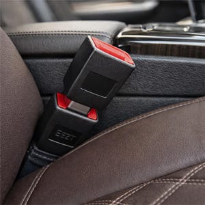 Op de afbeelding: Close-up van een zwarte veiligheidsgordelgesp met een rode ontgrendelingsknop. De gesp is verbonden met zijn tegenhanger, die het vergrendelingsmechanisme laat zien. De stoel is bruin leer met witte stiksels.