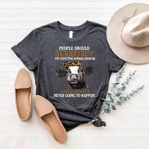May include: Dark gray t-shirt with a cow graphic wearing a leopard print bandana. The text on the shirt reads "PEOPLE SHOULD SERIOUSLY TOP EXPECTING NORMAL FROM ME WE ALL KNOW IT'S NEVER GOING TO HAPPEN!". Two pairs of tan flats and a beige hat are also in the image.