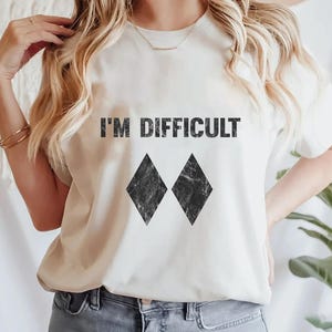 May include: A cream-coloured t-shirt featuring the text "I'M DIFFICULT" in black, with two black diamond shapes below. The t-shirt is a relaxed fit, ideal for casual wear.