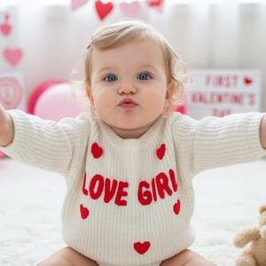 Peut inclure: Un pull en tricot à manches longues de couleur crème avec le texte rouge LOVE GIRL sur la poitrine, agrémenté de petits motifs de cœurs rouges dispersés sur le devant. Le pull présente un col rond côtelé et une maille texturée douce.