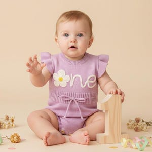 May include: A light purple knit romper featuring ruffled sleeves, a ribbed waist with a decorative tie, and an embroidered white flower next to the word one in white script. The garment has a round neckline and a buttoned bottom closure.