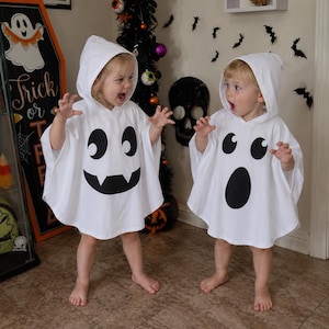 May include: Two white hooded ghost ponchos featuring black screen-printed facial expressions. One poncho displays a smiling ghost face with two fangs, while the other features a surprised ghost face with a large oval mouth and round eyes.