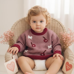 May include: A mauve long-sleeve knit sweater featuring a crew neckline, dark red ribbed cuffs and hem, and a pattern of embroidered white bows and dark red cherries. The sweater is worn over a white diaper.