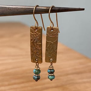 May include: A pair of brass earrings with a rectangular shape and floral pattern. Each earring has a gold-colored hook and is adorned with two turquoise-colored beads. The earrings are displayed on a metal tool.