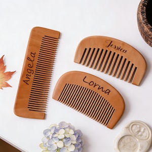 May include: Three wooden combs with personalized names: "Angela", "Jessica", and "Lorna". The combs are light brown with fine teeth. A decorative maple leaf and floral arrangement are also present.