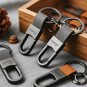 Puede incluir: Una colección de llaveros personalizados con correas de cuero gris y herrajes metálicos. Cada llavero presenta un nombre grabado en el cierre metálico. Un llavero tiene una correa de cuero marrón. Los llaveros están colocados cerca de una caja de madera y una piña.