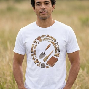 May include: White t-shirt with a circular design of bones, a small shovel, a brush, and a book in shades of brown and beige. The shirt is worn by a person standing in a field of tall grass. The design has a natural history theme.